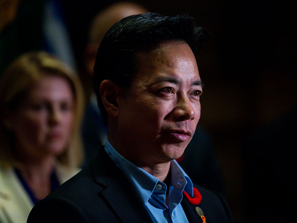 A close-up photo of Ken Sim at a press conference, wearing a red poppy on his lapel. Some other people are visible behind him, but blurry. 