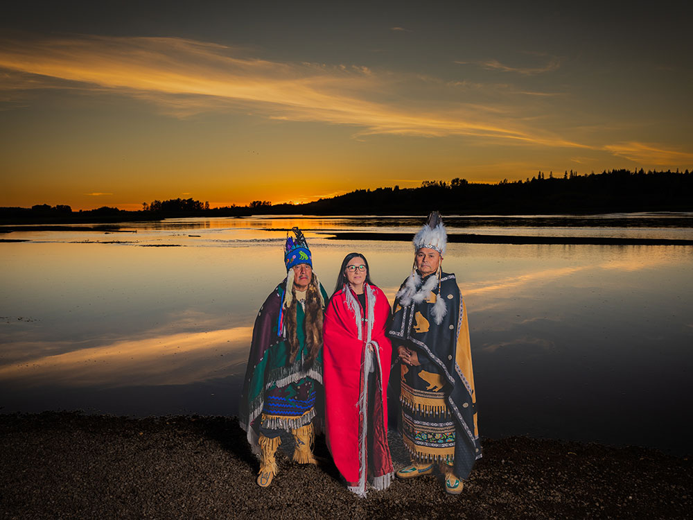 An Indigenous woman, flanked by two men, stands on the ban of a river at sunset. The water is calm and the sky is blue and gold, with clouds. All three are wearing regalia.