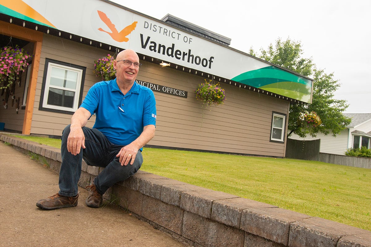 A smiling man in a blue shirt and jeans sits on a low retaining wall in front of the Vanderhoof municipal offices.