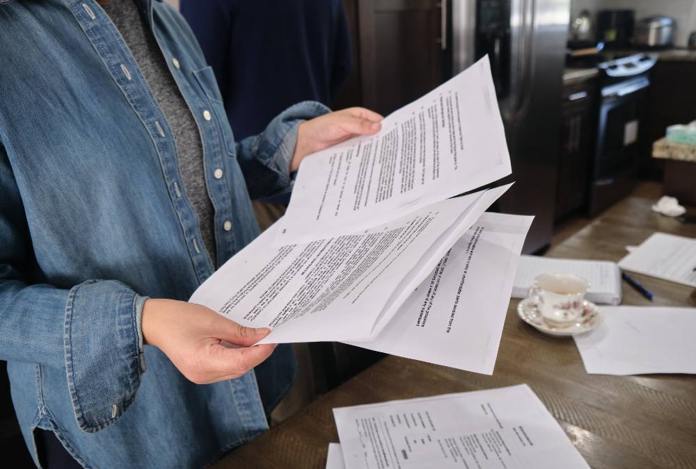 A woman holds several black and white computer printouts in her hands at a brown kitchen table. She is wearing a denim button-down shirt over a grey t-shirt. A cup of tea sits on the counter and a stainless-steel refrigerator is visible in the background. 