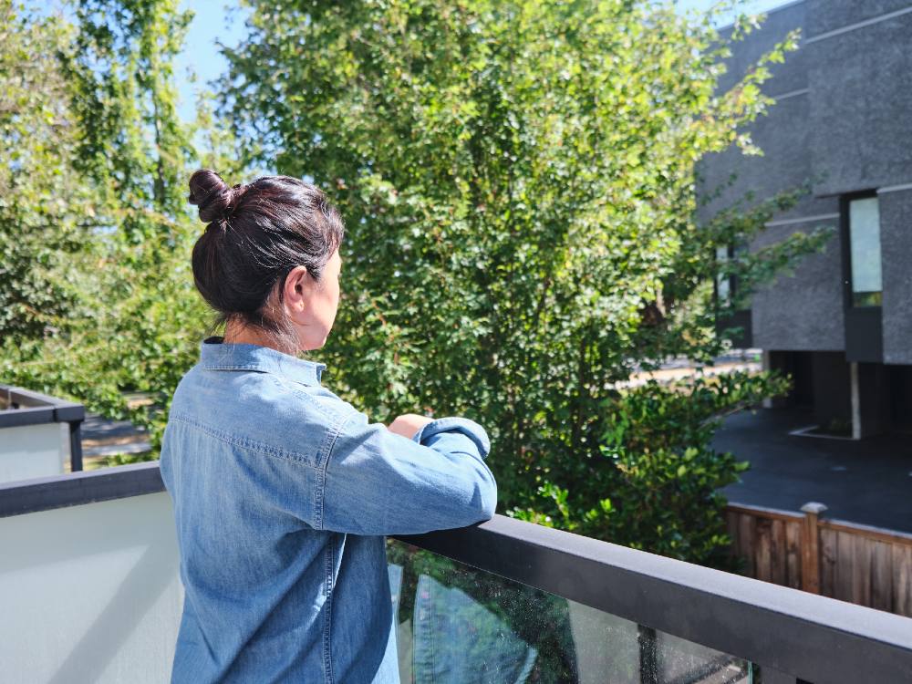 A Chinese woman stands on a balcony overlooking a crop of trees and a neighbouring grey building. It’s a sunny day. The woman is turned away from the camera and she is resting her arms on the balcony ledge. She is wearing a denim shirt and her black hair is tied in a topknot bun.