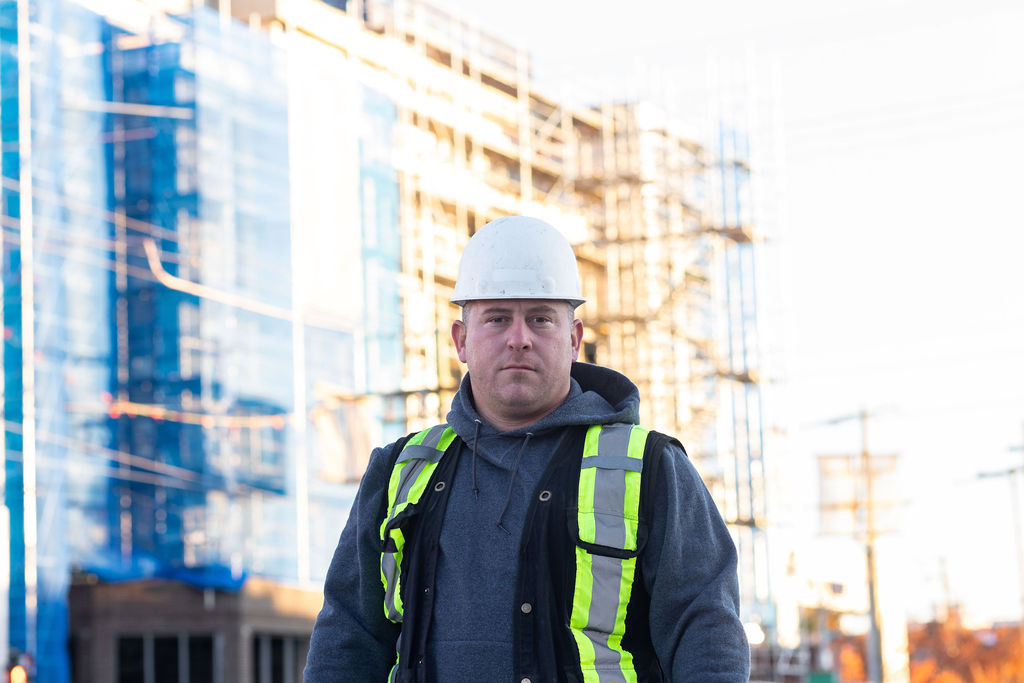 A man wearing a white hardhat, grey hoodie and reflective safety vest looks at the camera. In the background is a multi-storey building under construction.