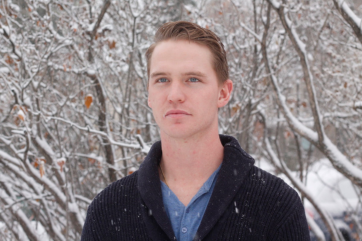 A clean-shaven man with short brown hair looks at the camera. He’s wearing a black sweater with a collar and blue shirt. In the background are small, snow-covered trees.