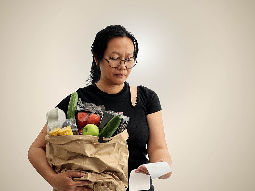 A woman holding a paper bag of groceries is looking down at a receipt.