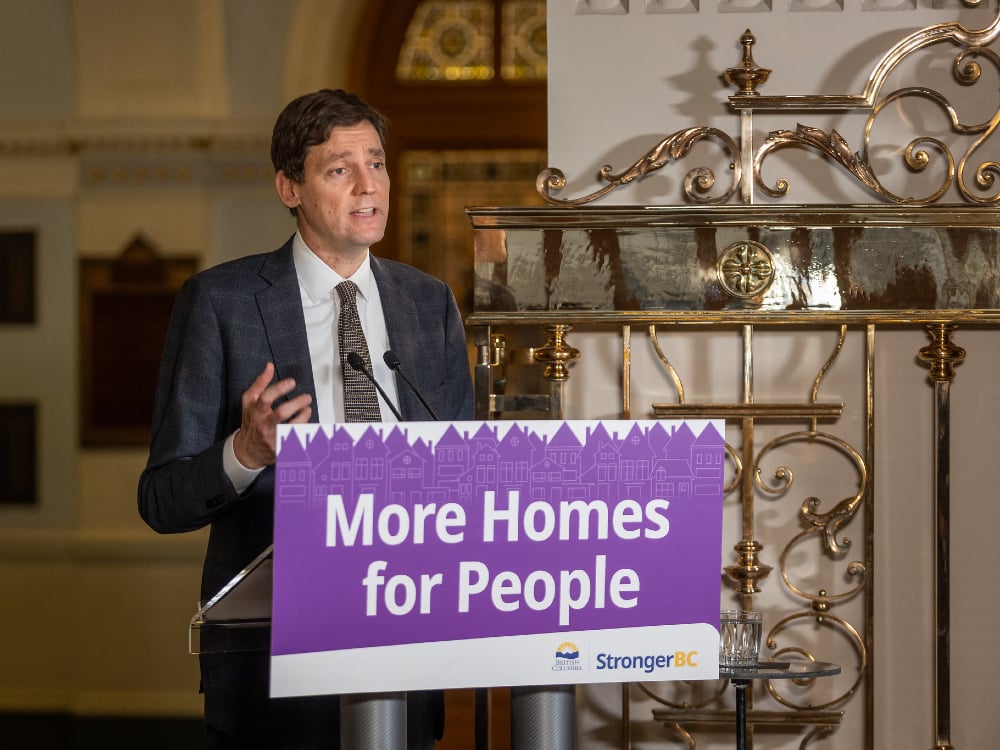 Premier David Eby stands at a podium. A purple sign on the podium reads, ‘More Homes for People.’