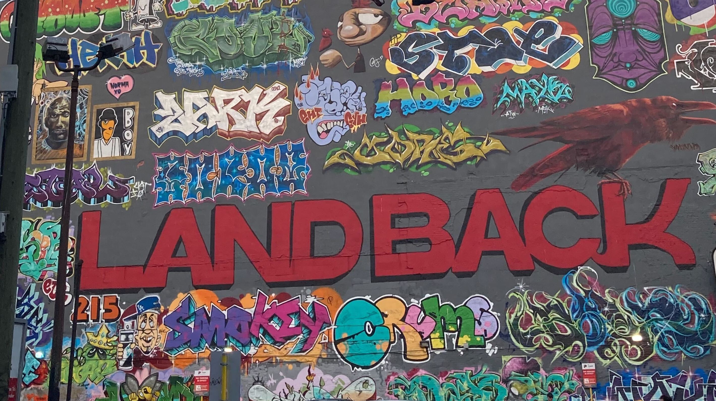 A large-format graffiti mural features numerous colourful tags from local artists unified by the words “LAND BACK” in large sans-serif lettering in red. The main lettering and surrounding colourful tags are on a grey charcoal painted wall.