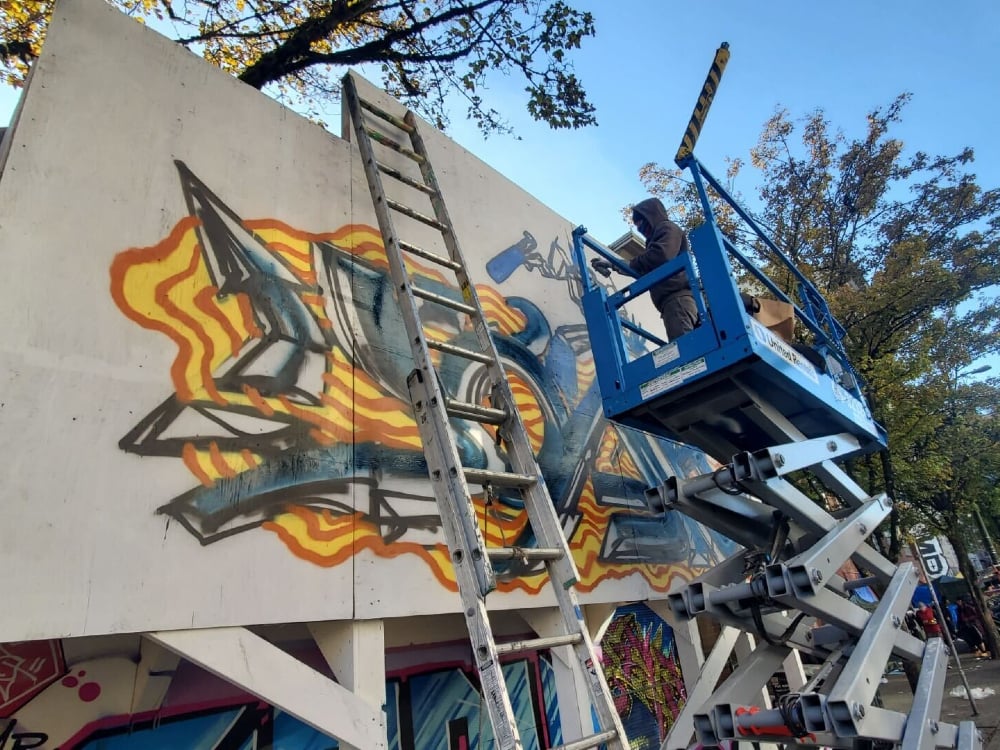 A man in a dark grey hoodie over a tuque and mask stands in a blue cherry picker while he sprays paint on a graffiti wall in Vancouver’s Downtown Eastside. The artwork uses orange, yellow, blue and black paint. A silver ladder is in the foreground against the wall. In the background is a stand of green trees and a busy sidewalk full of people.
