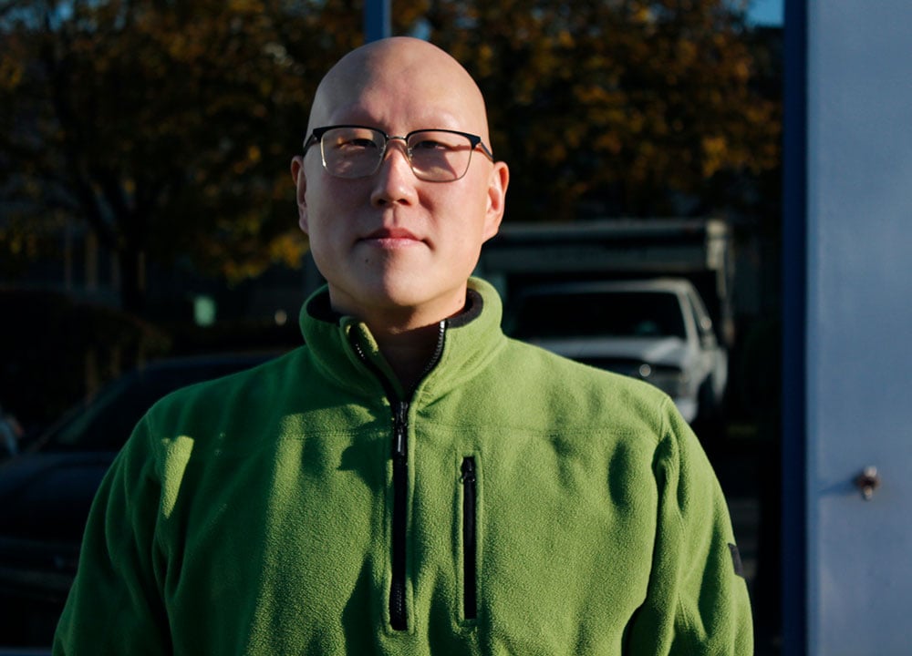 A man in a light green fleece jacket, with glasses and no hair, looks at the camera. It’s outdoors, and sun illuminates the right side of his face.