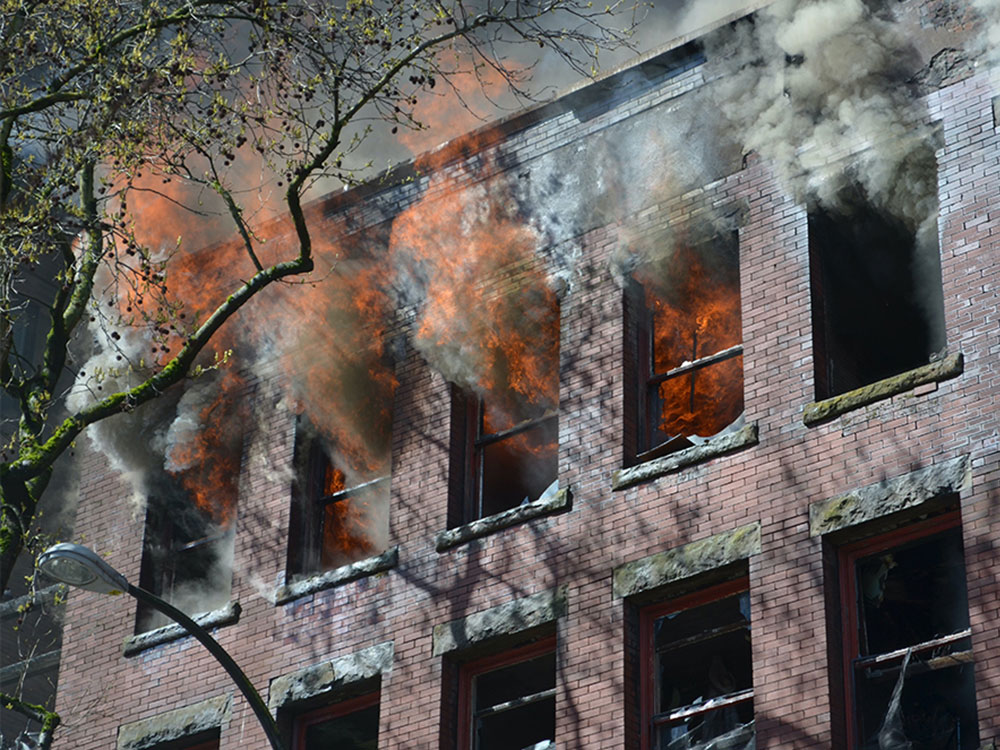 The upper floor of an older red-brick building, with a series of windows. Flames are coming out of some windows; smoke from others.