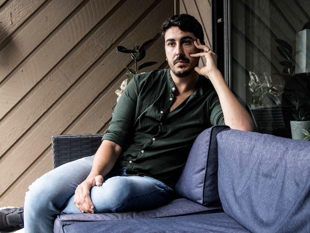 A man sits on a couch on a balcony, resting part of the weight of his head on his hand. His gaze does not meet the camera.