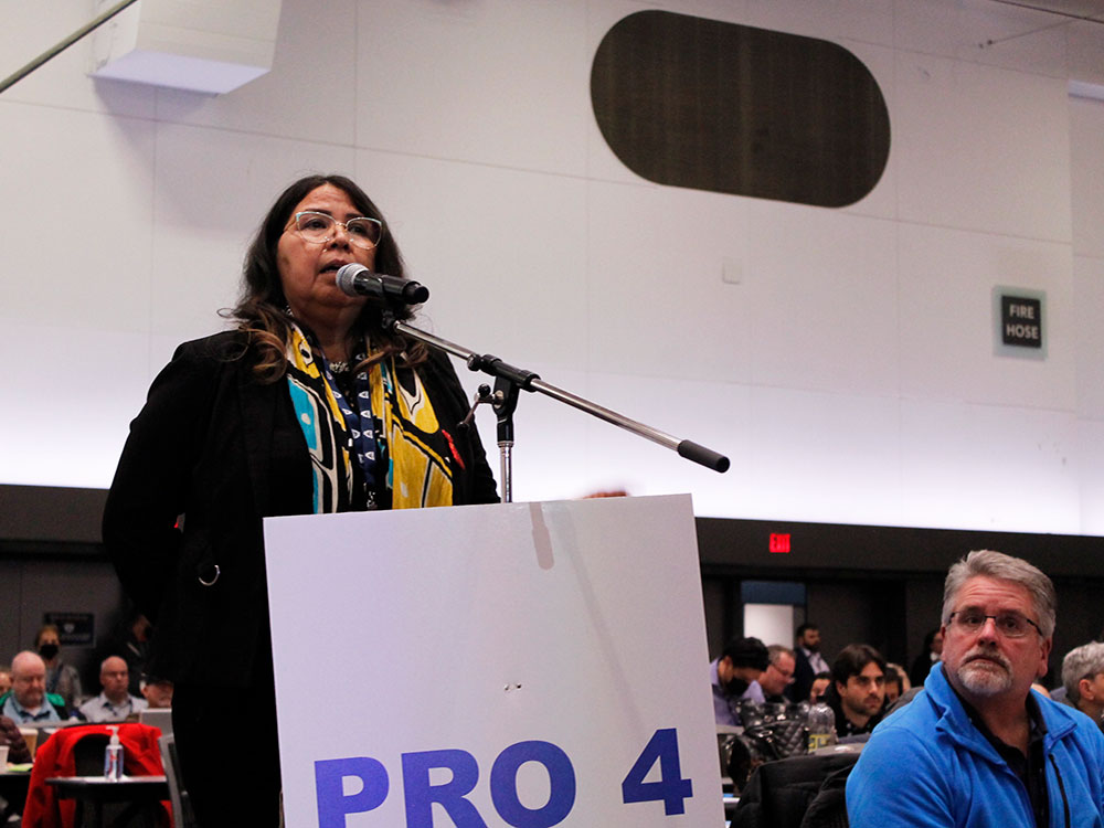 A woman stands at a podium, addressing a number of people at a conference.