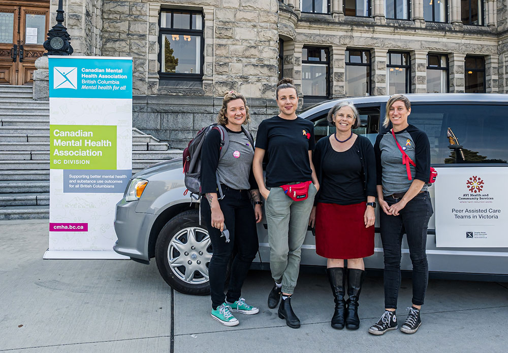 Four people stand in front of an SUV parked in front of the provincial legislature in Victoria. A large sign reads, ‘Canadian Mental Health Association, BC Division.’