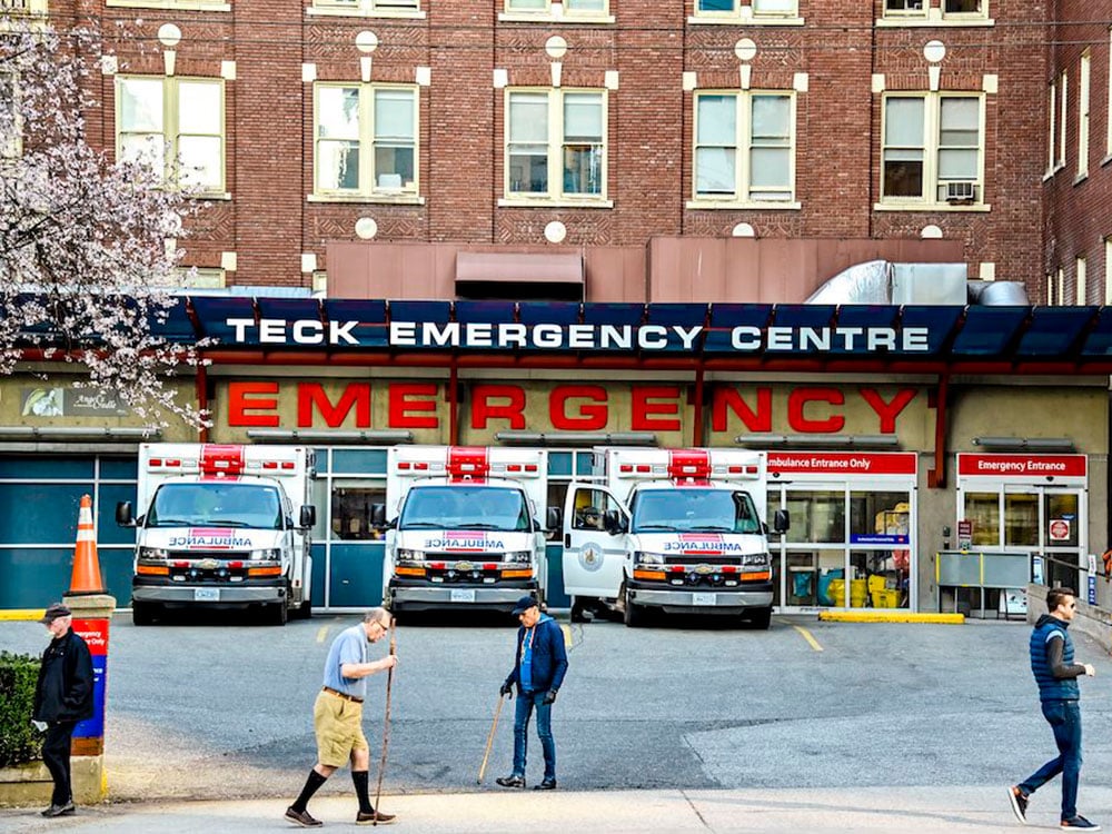 City dwellers stroll in front of a concrete and glass hospital entrance with three ambulances parked and large signs saying Emergency in red and Teck Emergency Centre in white. 