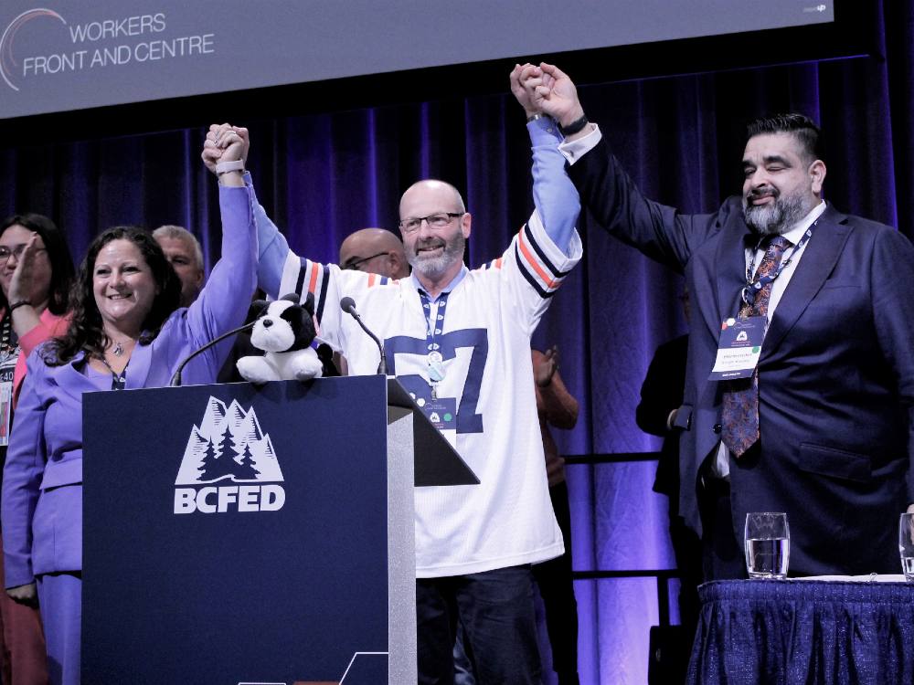 A woman with long dark hair wearing a blue jacket stands beside a man wearing glasses and sports sweater. On his left is a man in a blue suit, white short and tie. All three are raising clasped hands and standing behind a BC Fed logo. 