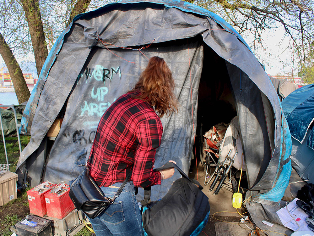 A woman with her back to the camera stands outside of a tent in Crab Park. Tents and belongings can be seen in the background.