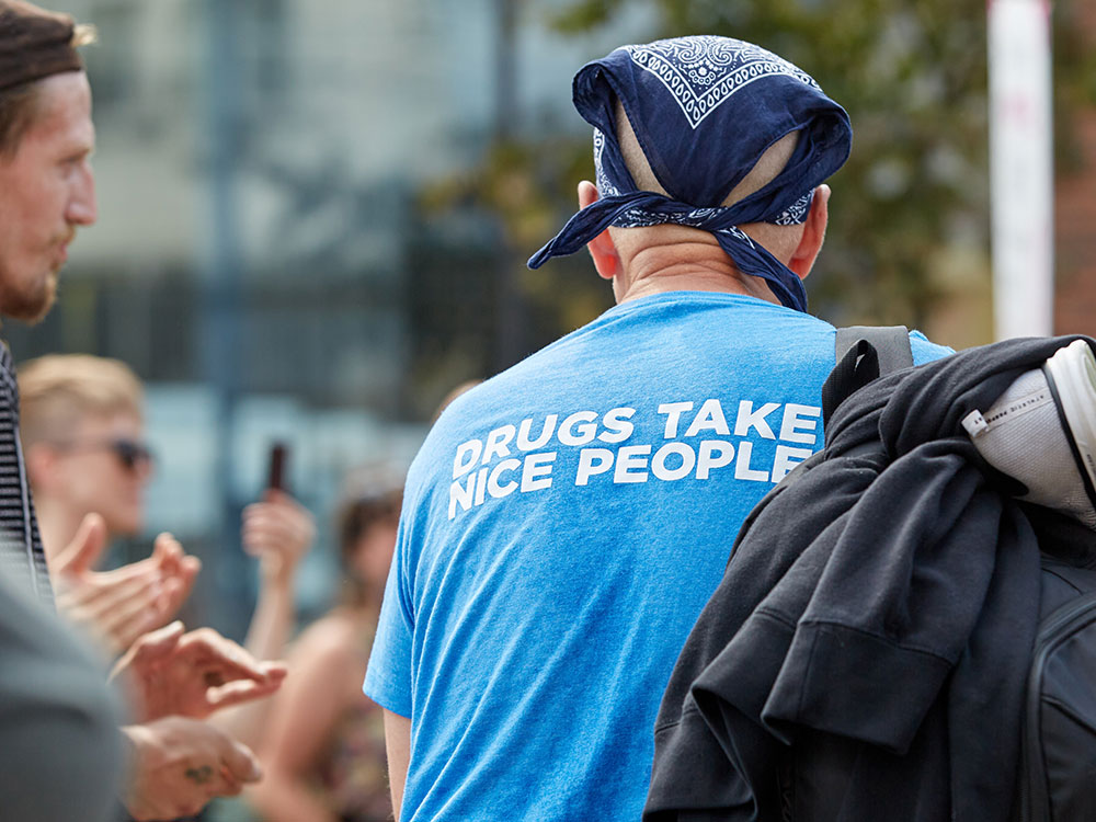 A man, viewed from behind, wears a blue bandanna, a backpack, and a T-shirt that reads, “Drugs Take Nice People.”