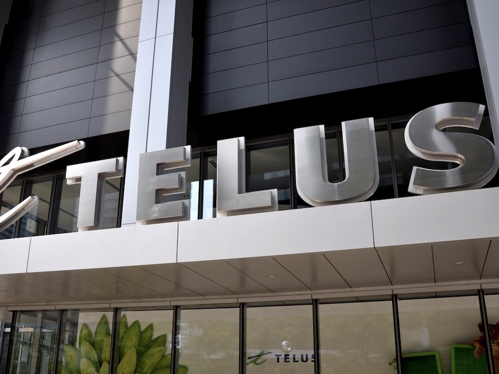 The photo shows an urban office building with a glass-walled lobby. Above the door silver sculpted letters spell out TELUS.