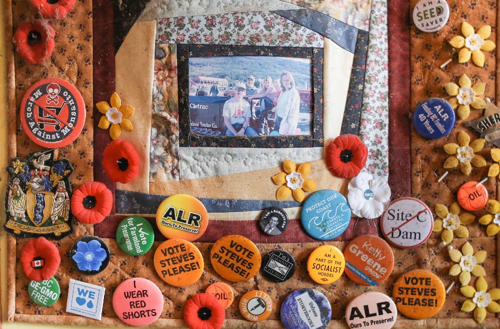 Colourful circular political buttons, many of them orange and blue hang on an orange quilt on the wall with a patch of an old colour family photo in the centre. Some of them read: “Vote Steves please!” “No Site C Dam.” “I wear read shorts.” “I am part of the socialist hordes.” “ALR: ours to preserve!” 