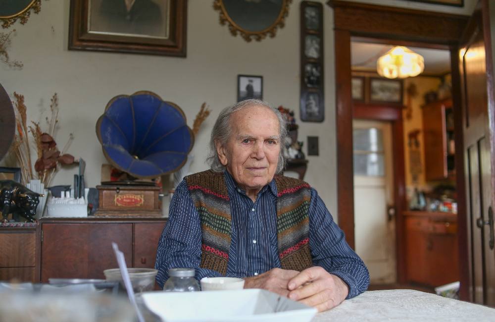 An 85-year-old man sits at a dining table, looking at the camera. He’s wearing a pinstriped blue collared with a striped sweater vest with buttons. Behind him are old pictures and a blue phonograph. He’s in a cozy looking home with worn wood beams and furnishings.
