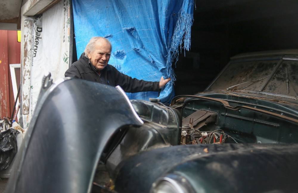 An 85-year-old man pulls aside a blue tarp to reveal a navy blue 1950 Dodge convertible that’s being fixed up. The hood is missing and the parts inside are visible.