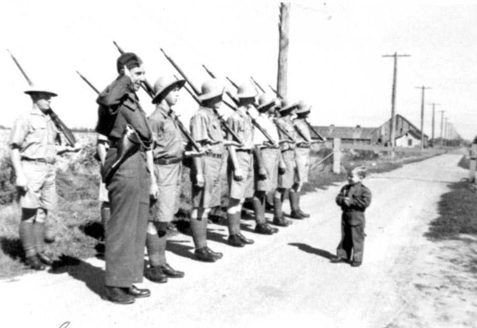 A black-and-white photo of a uniformed toddler inspecting a line of troops on a rural-looking road saluting him.