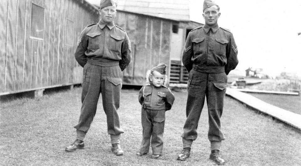 A black-and-white photo from the Second World War of a uniformed toddler between two soldiers.