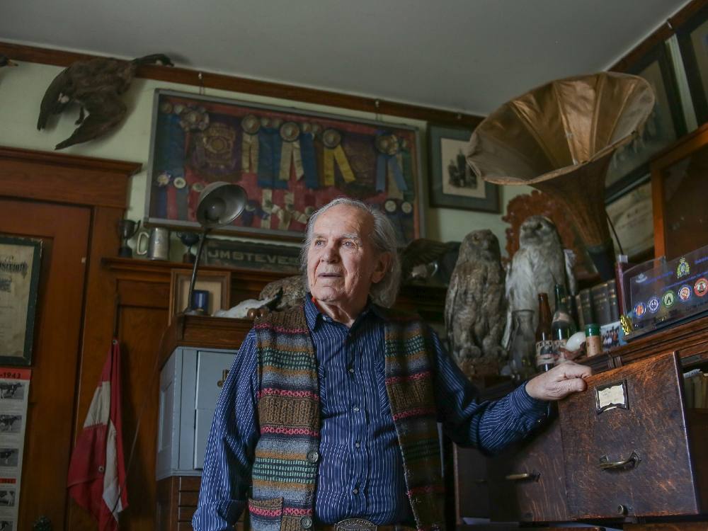 An 85-year-old man is standing in the middle of a room. He’s wearing a blue pinstriped shirt under a knitted vest in brown, pink and blue. He has longish, wispy grey hair. He’s leaning on an aged, dark brown wooden cabinet. The room is filled with old memorabilia: taxidermy of ducks and owls; framed ribbons; and a large bronze phonograph.