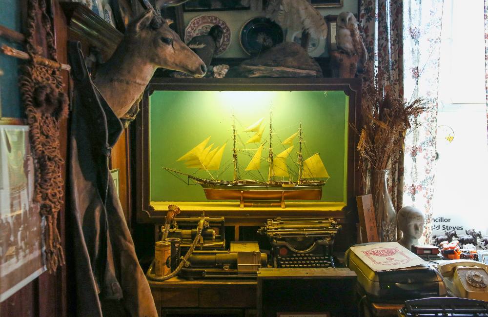 A model ship in a light-up display case. It is surrounded by other memorabilia including a mounted deer’s head and a typewriter.