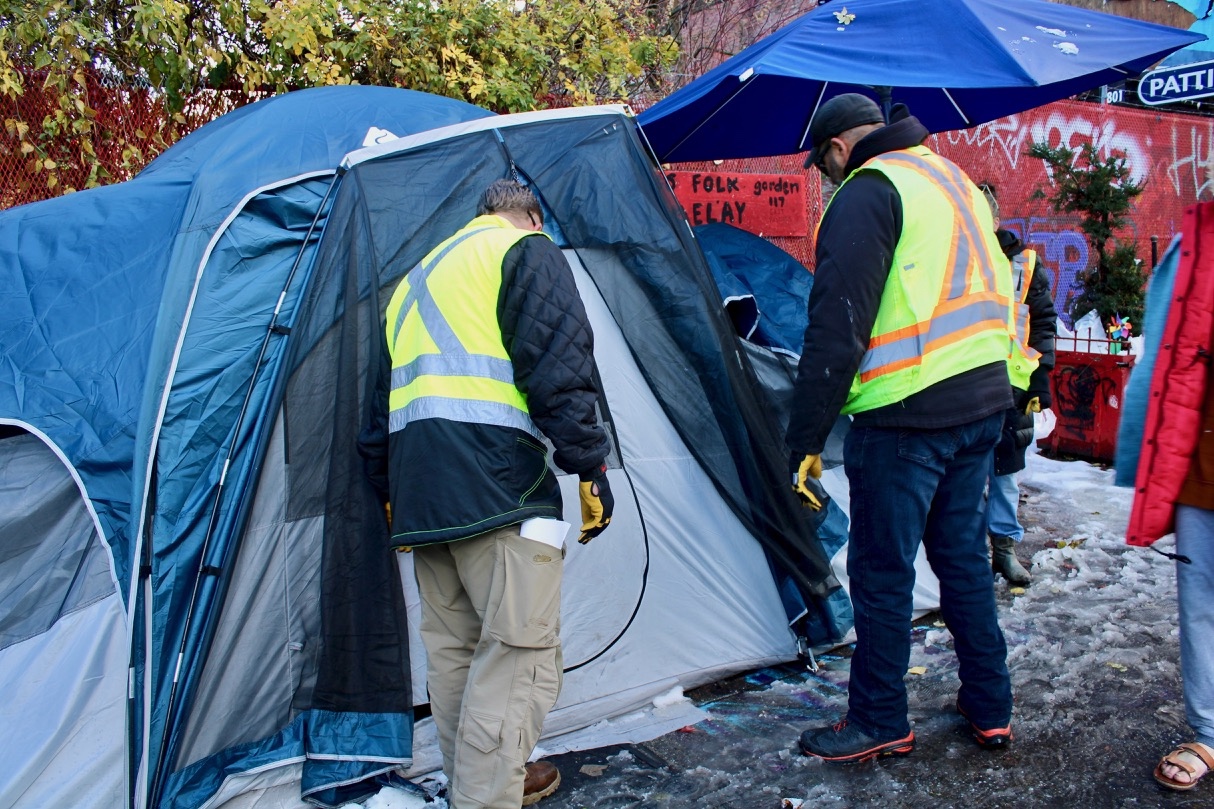 Two men in yellow reflective safety vests stand on an icy sidewalk outside a blue tent.