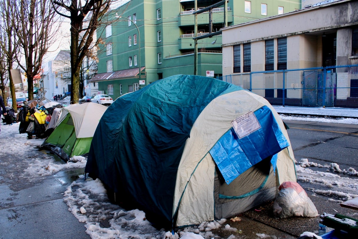 A row of tents lines the outer edge of a snowy sidewalk on a bleak street. The first tent has a printed notice hanging over the door.