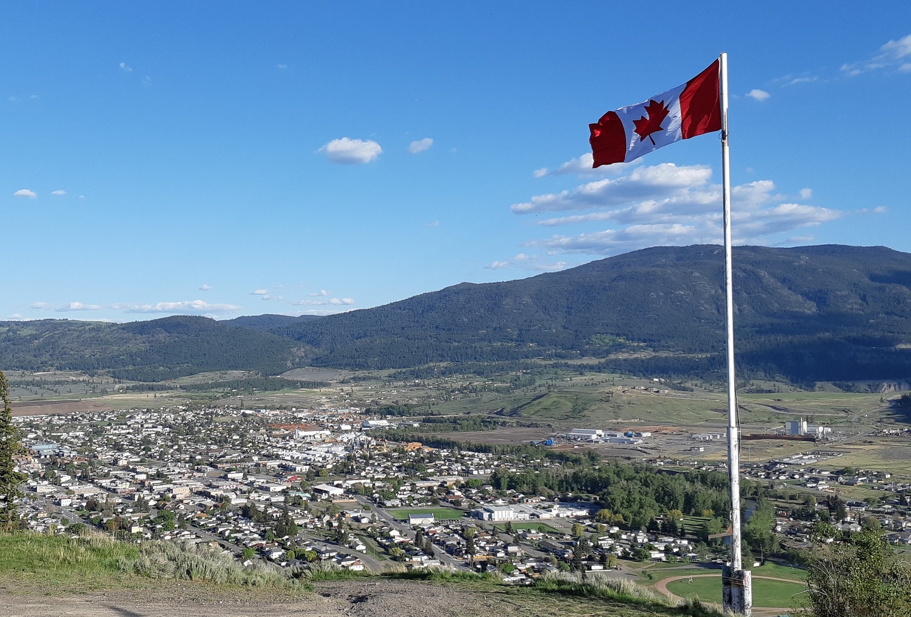 A Canadian flag flies above a flat town in the middle of a valley. There’s a clear blue sky.