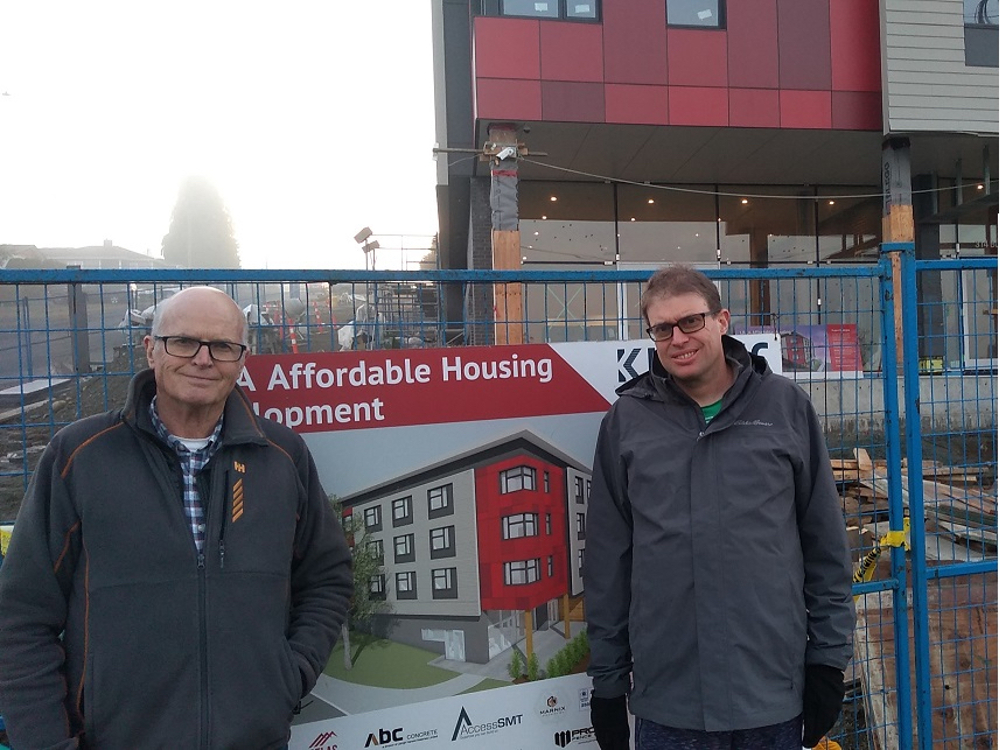 Two people in grey jackets stand in front of a sign announcing a new affordable housing project. The building is behind them.