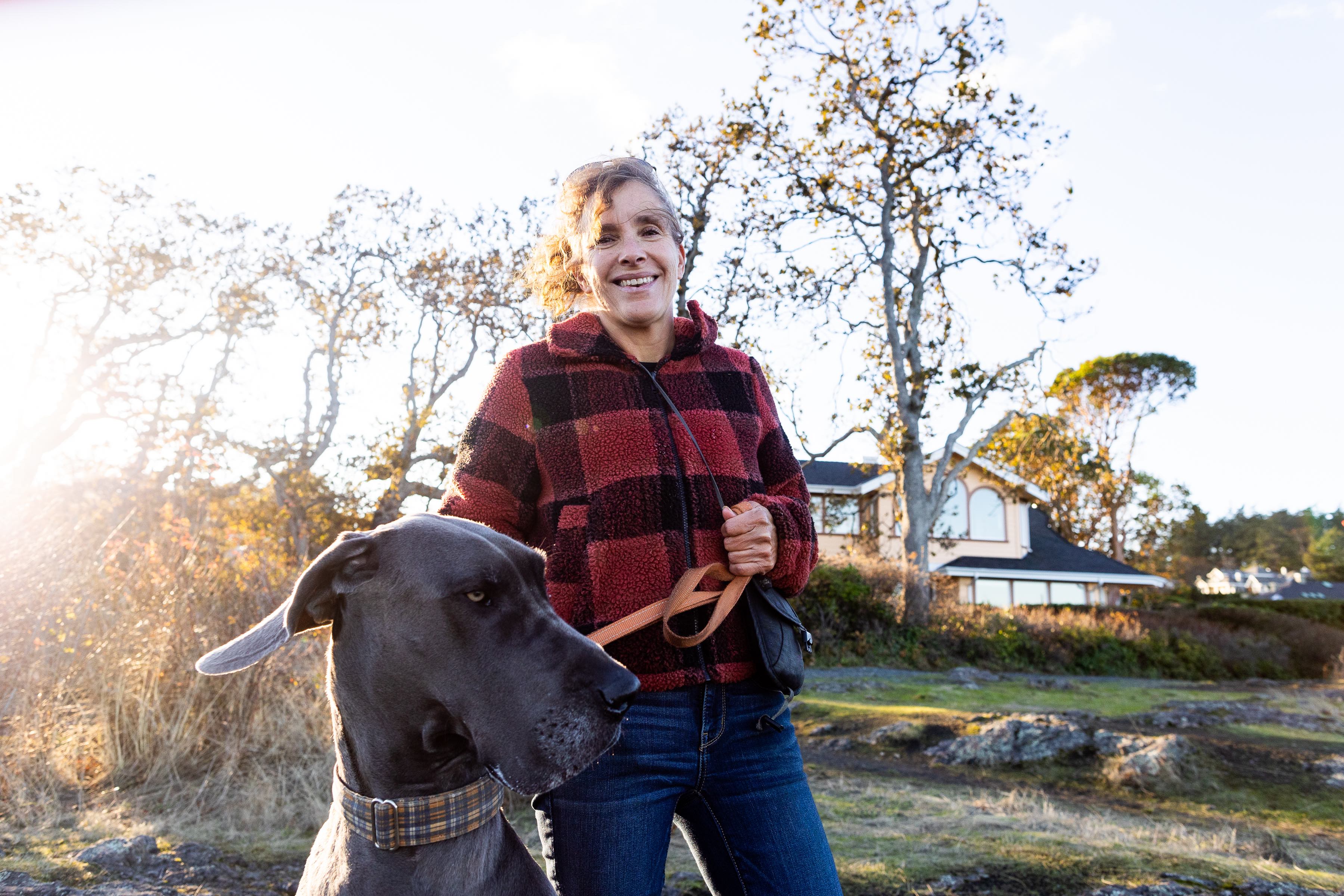 A woman wearing a red and black fleece jacket and jeans smiles at the camera. She is reaching down to pat a Great Dane and holding a smaller dog. In the background, waves crash on rocks.