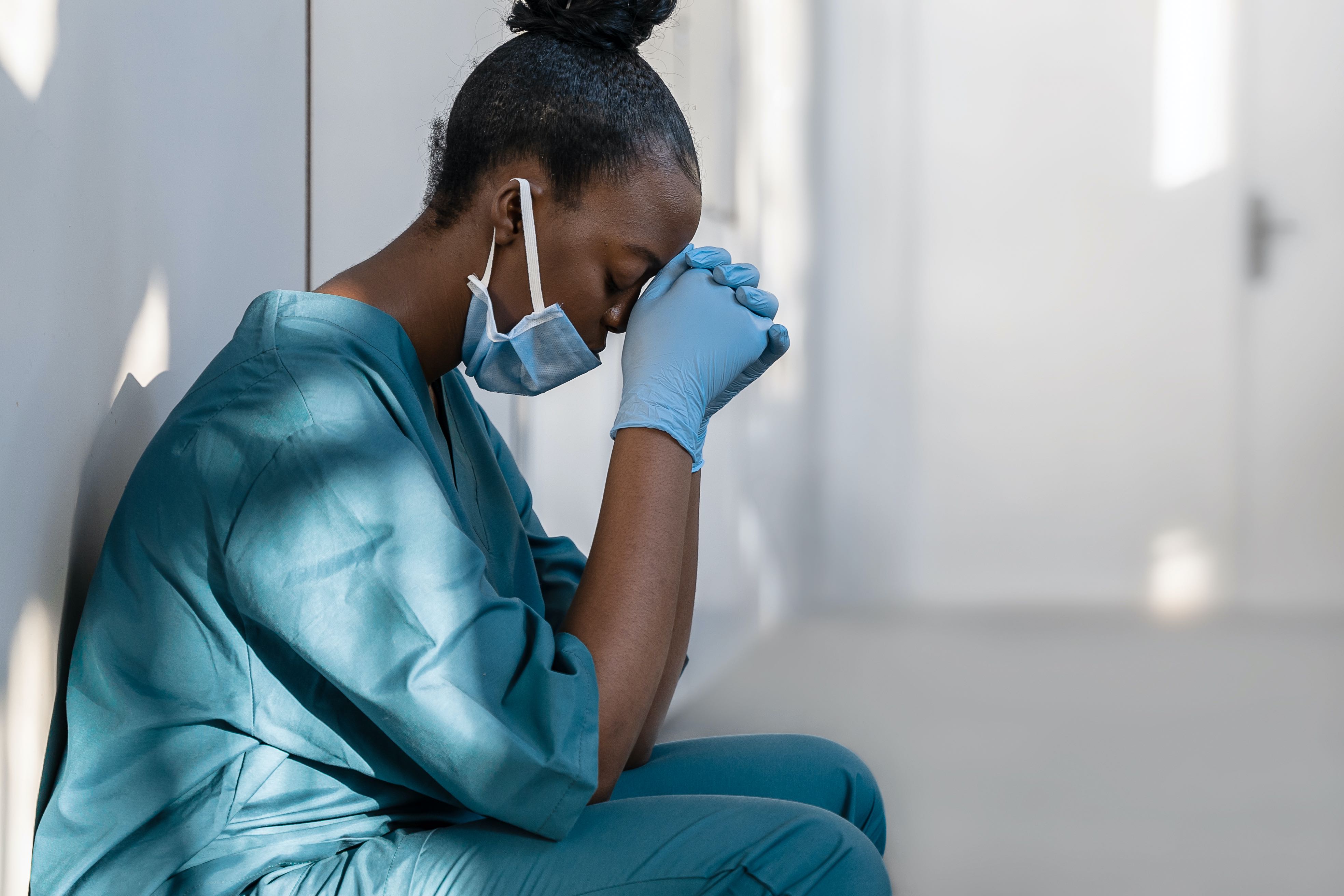 A woman in a hospital uniform and a mask sits against a wall. Her eyes are closed and she appears exhausted.