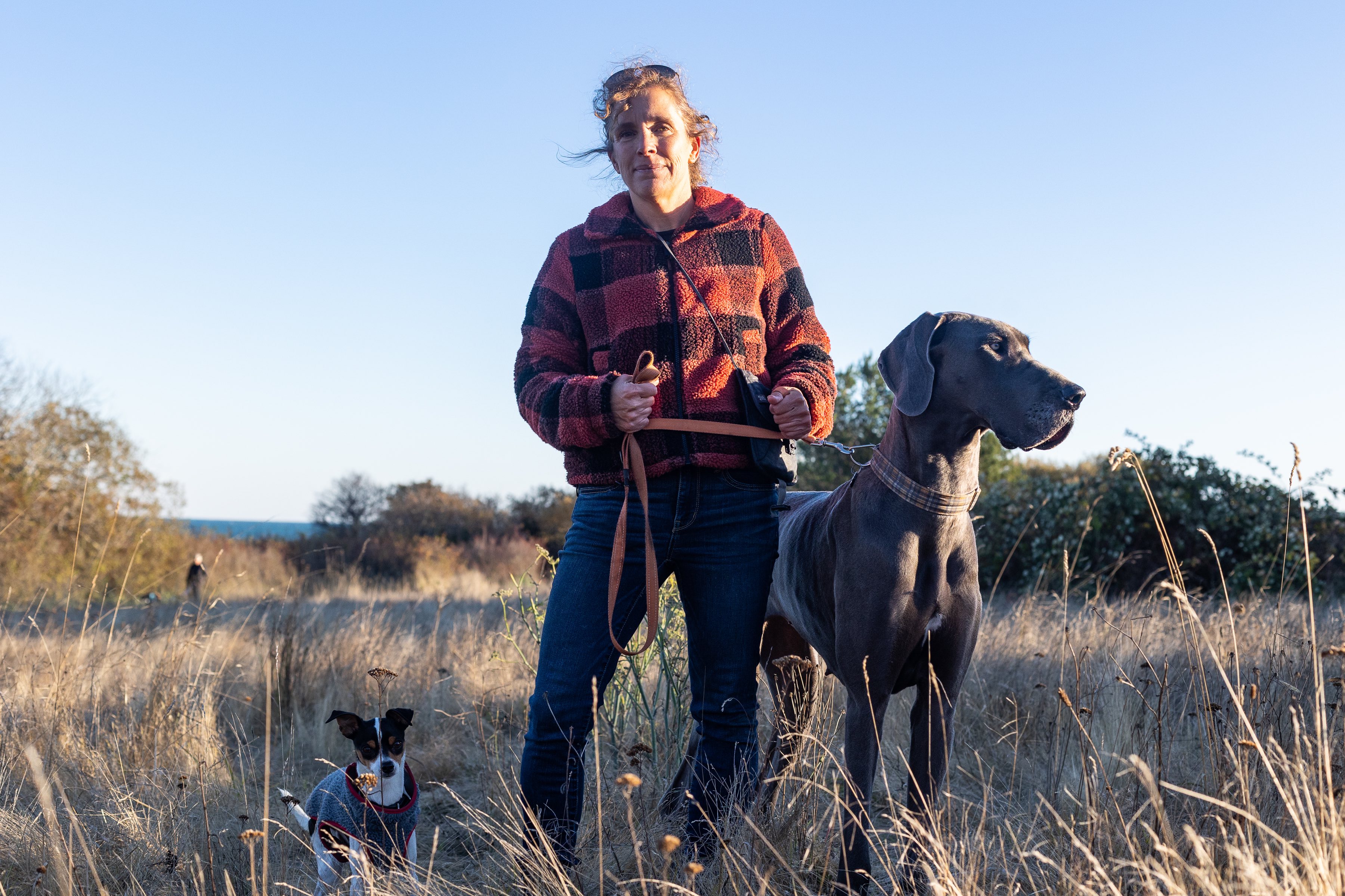 A woman wearing a red and black fleece jacket and jeans holds a Great Dane on a leash. A smaller dog is beside her. She stands in a meadow of brown grass, with the ocean in the background.