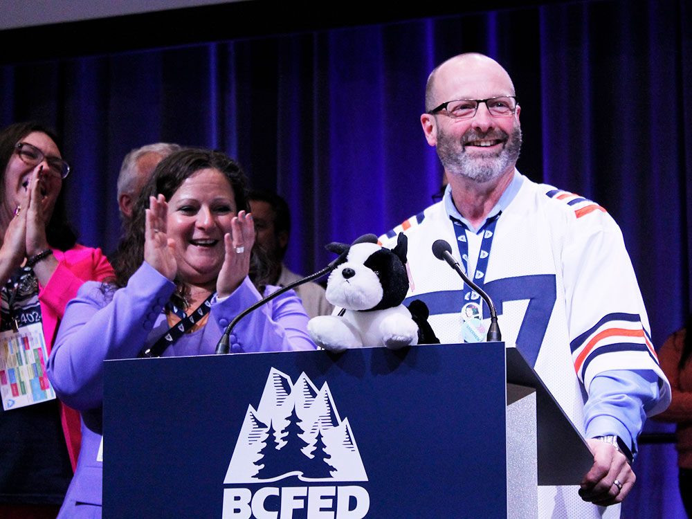 A woman in a lilac suit claps her hands. Beside her, a man wearing a sports jersey, with a neatly trimmed beard and wearing glasses, smiles. They stand behind a podium with a blue sign saying BC Fed.