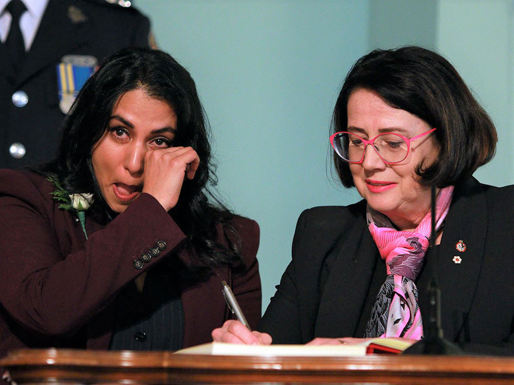 A black-haired woman wearing a burgundy suit wipes a tear from her eye. Another woman, wearing a black suit, a scarf and red glasses, sits beside her.