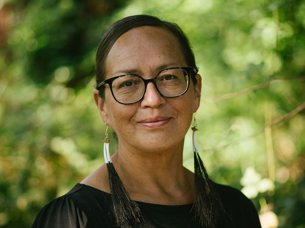 A woman wearing glasses and long earrings looks at the camera with a slight smile.
