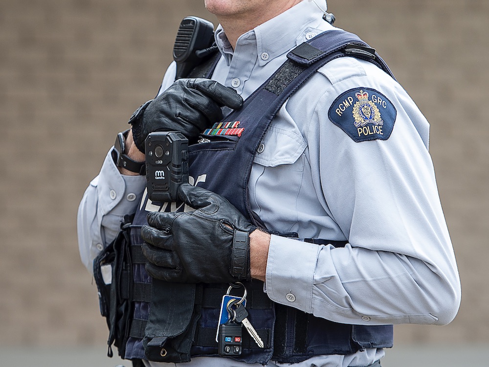 A male RCMP officer is wearing a uniform and a black stab vest with a microphone and other equipment. A video camera is attached to his chest. Only his torso is shown.