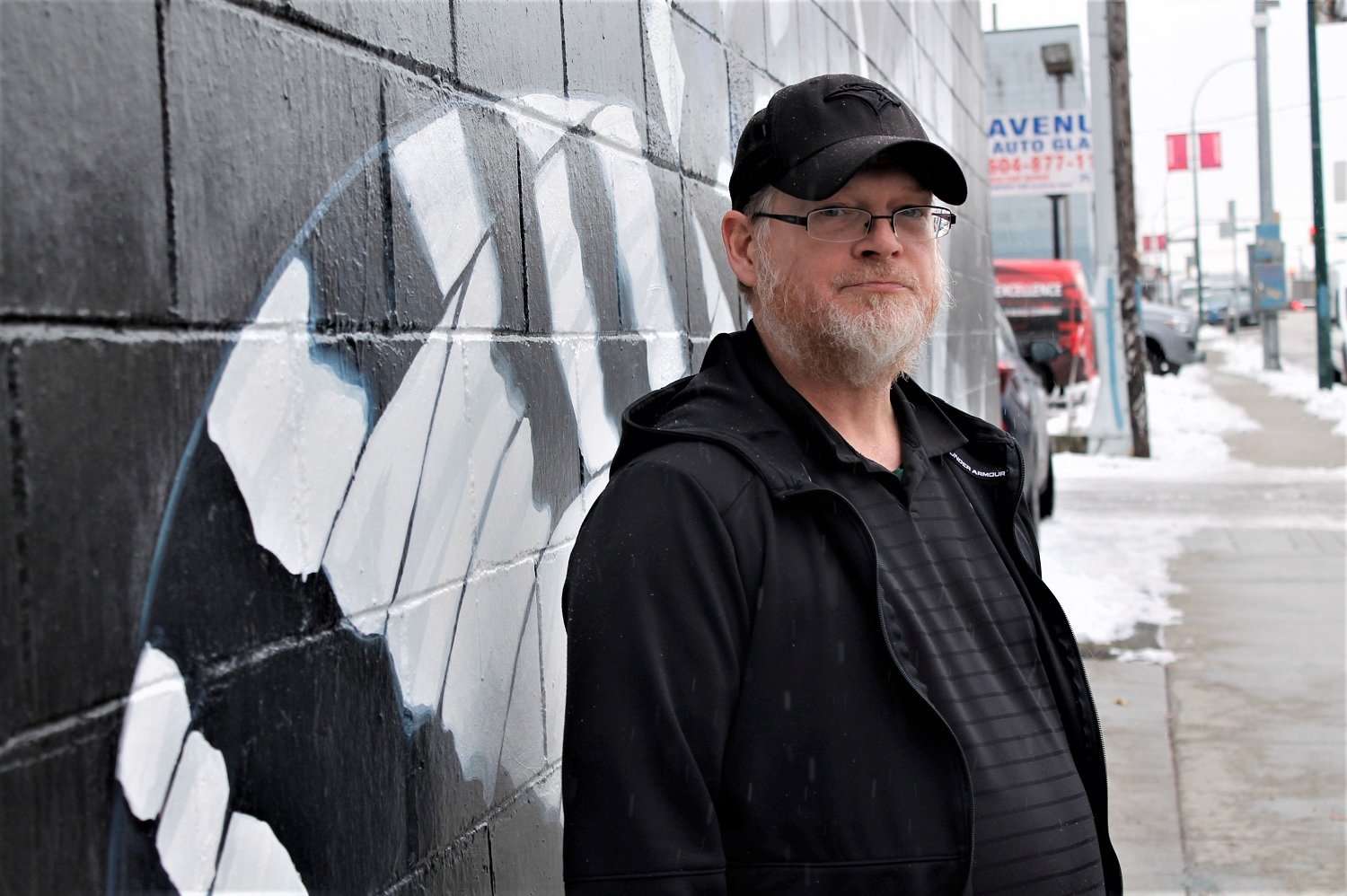 A man wearing a black hoodie and a black ballcap stands on the snowy sidewalk next to a black-and-white mural.
