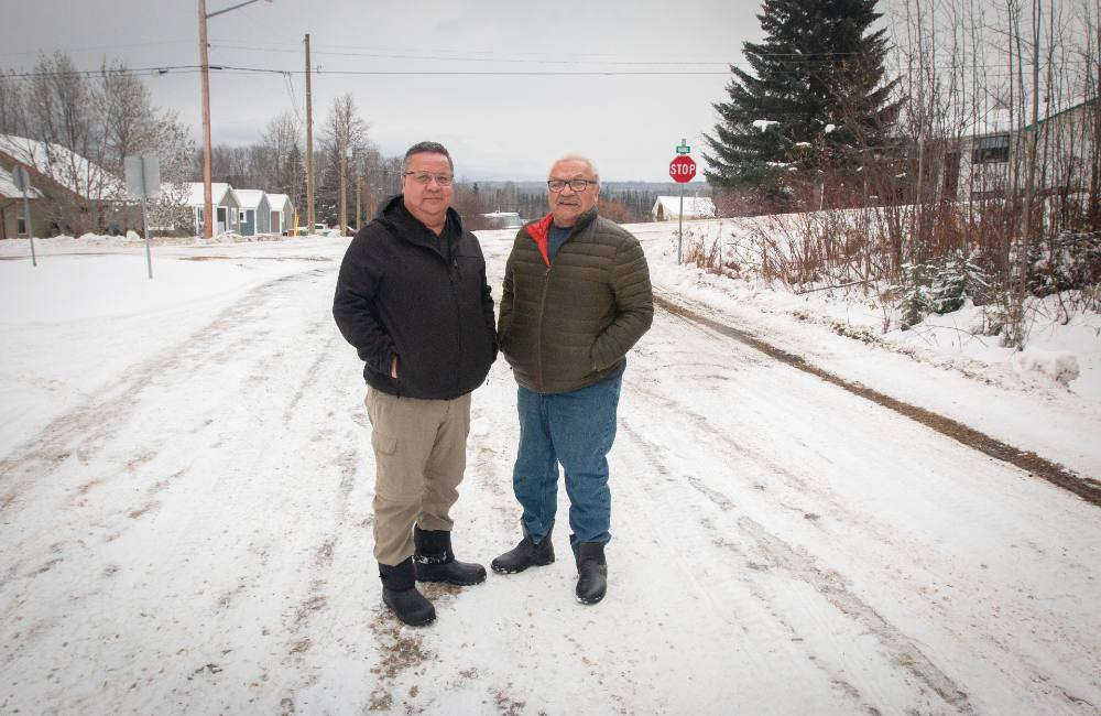 Two men stand on a snowy road. The man on the left wears a black coat and beige pants. The man on the right wears a brown down jacket and jeans. 