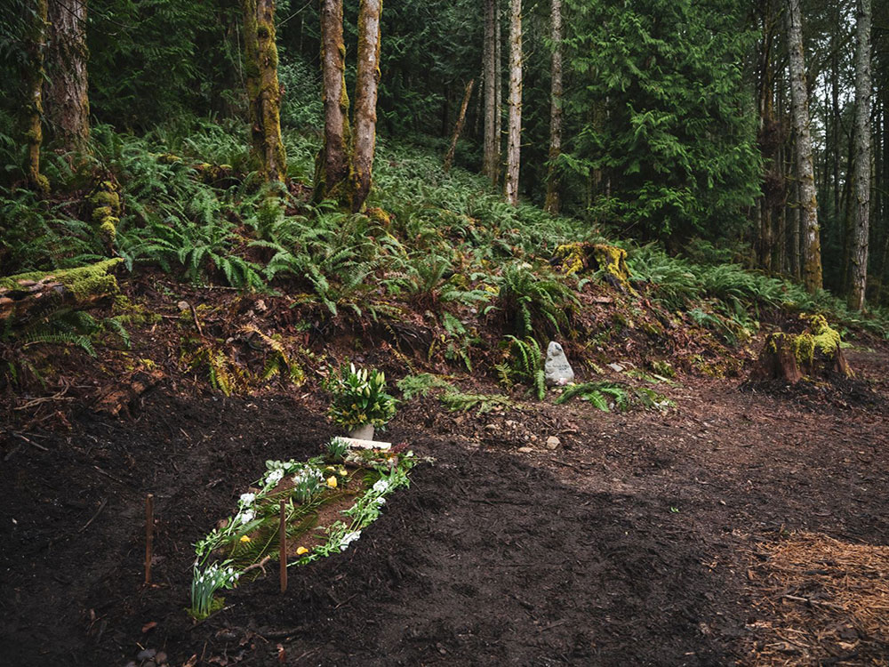 In a wooded West Coast forest a fresh grave has been covered with dirt. White flowers are arranged around it. In the background you can see sword ferns and cedar trees which are characteristics of a temperate coastal rainforest. 