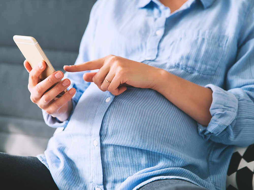 A pregnant person in a blue button-down shirt sitting on a grey sofa types into a white smartphone with one hand while holding it in the other.