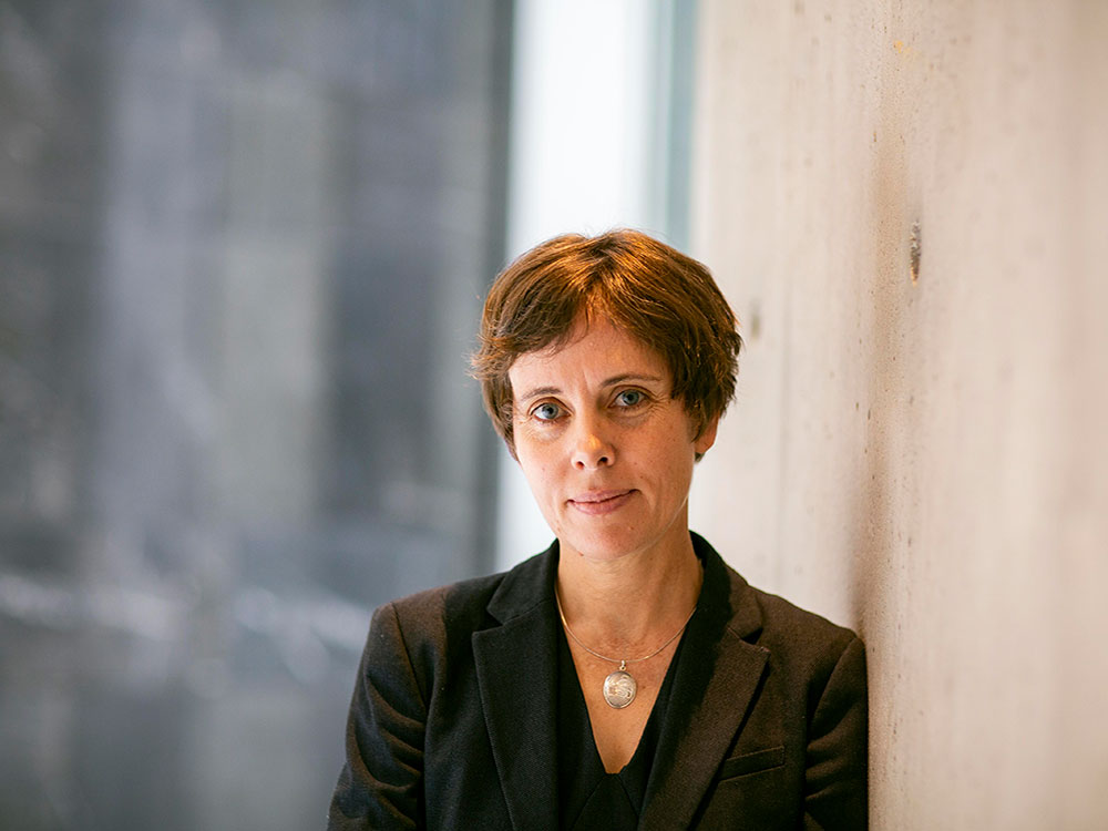 A woman with short brown hair looks at the camera. She is wearing a black shirt and black suit jacket, with a silver necklace.