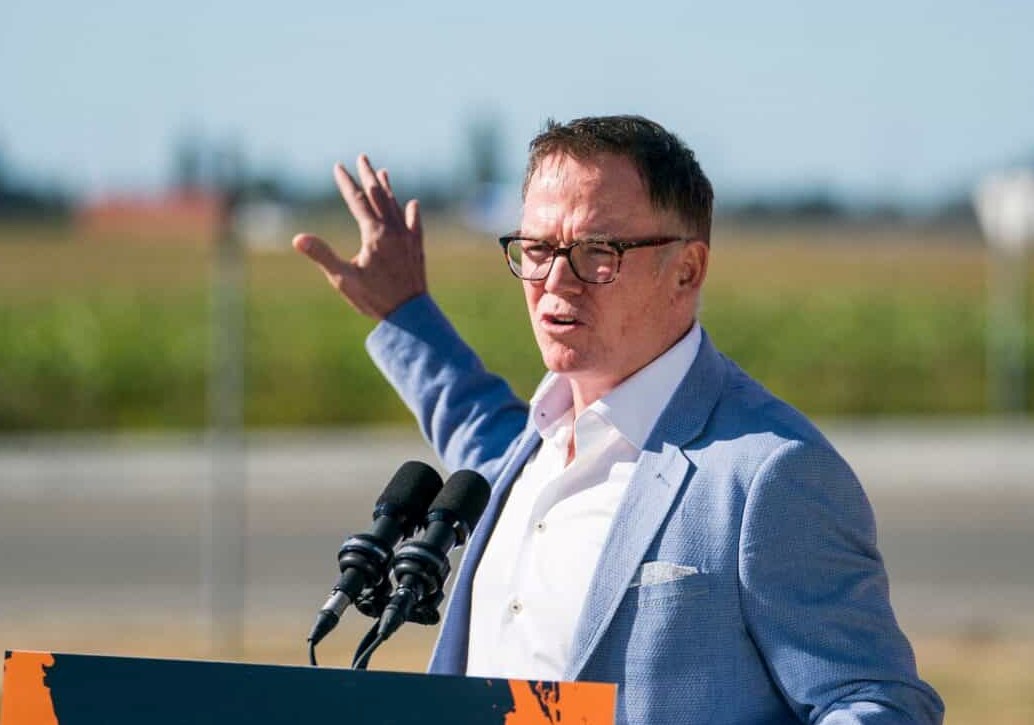 Kevin Falcon, wearing a light blue jacket and white shirt, gestures from behind a podium.