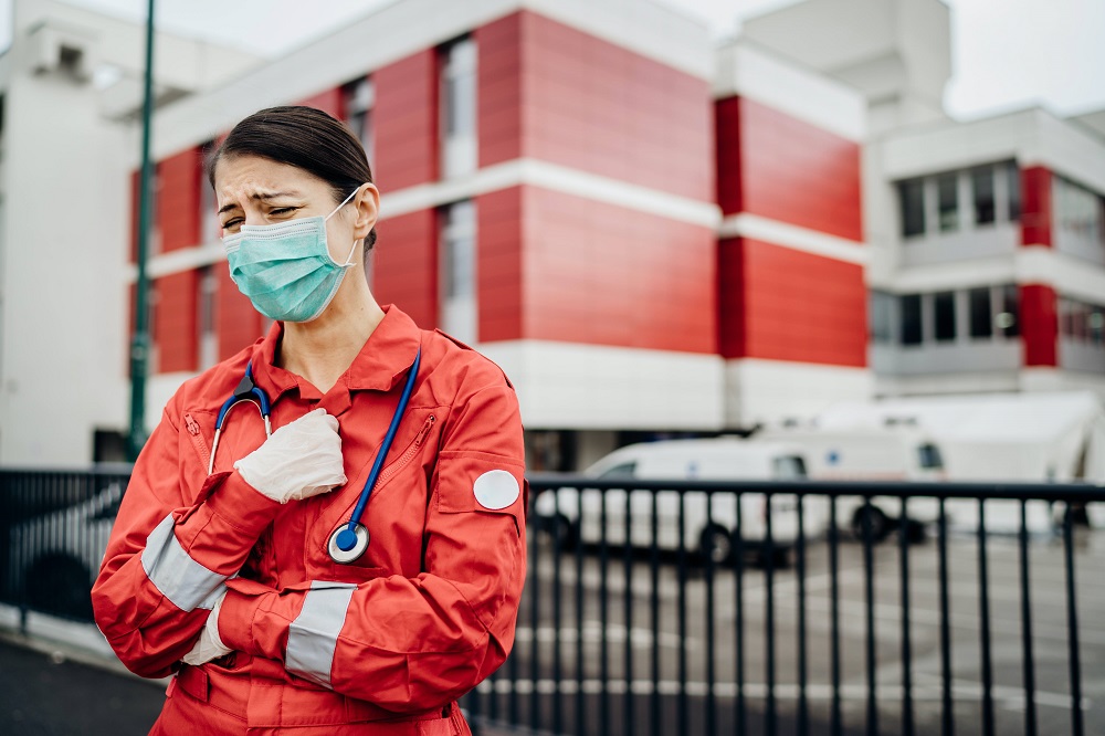 A woman in red scrubs with a stethoscope draped around her neck, wearing a mask, stands in front of a hospital. She appears to be crying.