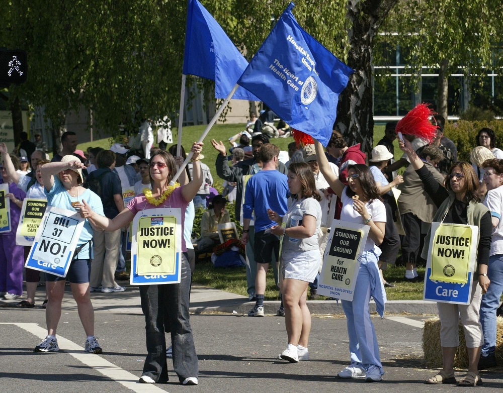 A group of women stand on the street on a summer day, some waving blue Hospital Employees’ Union flags and wearing placards that say “Justice Now” and “Standing Our Ground for Health Care.”