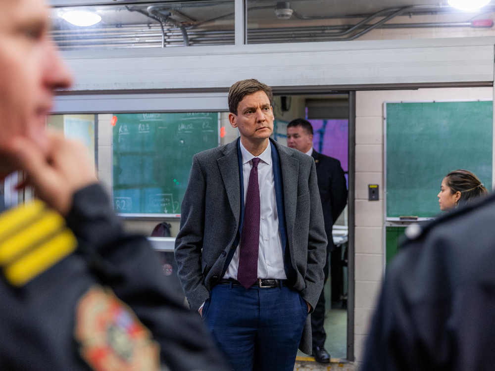 David Eby wears a blue suit, maroon tie and grey overcoat. He stands with his hands in his pockets and has a pensive look on his face. He appears to be in a police station.