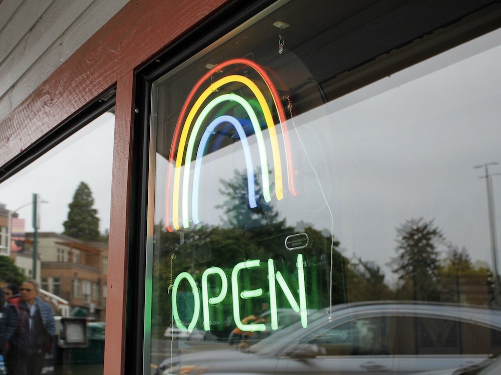 A storefront window, with a neon ‘OPEN’ sign under a Pride rainbow symbol. Pedestrians and cars are reflected in the window.