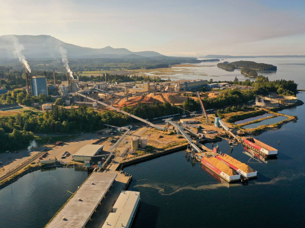 A bird’s eye view of the Crofton pulp and paper mill, surrounded by trees and the sea. In the foreground are piles of woodchips awaiting processing in large industrial structures left, two plumes of white gas rise from tall chutes to the left. 